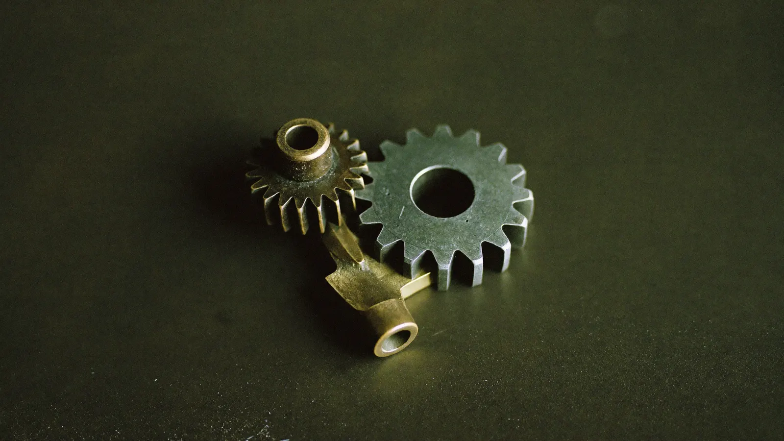 Two machined gears on a dark surface, one bronze and one green, cut to different pitches and not meshing.
